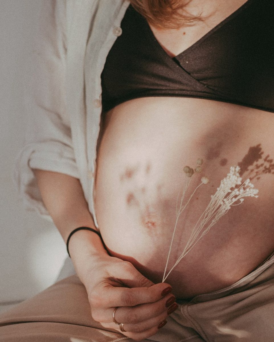 A pregnant belly in soft neutral light, a sprig of dried flowers held against it