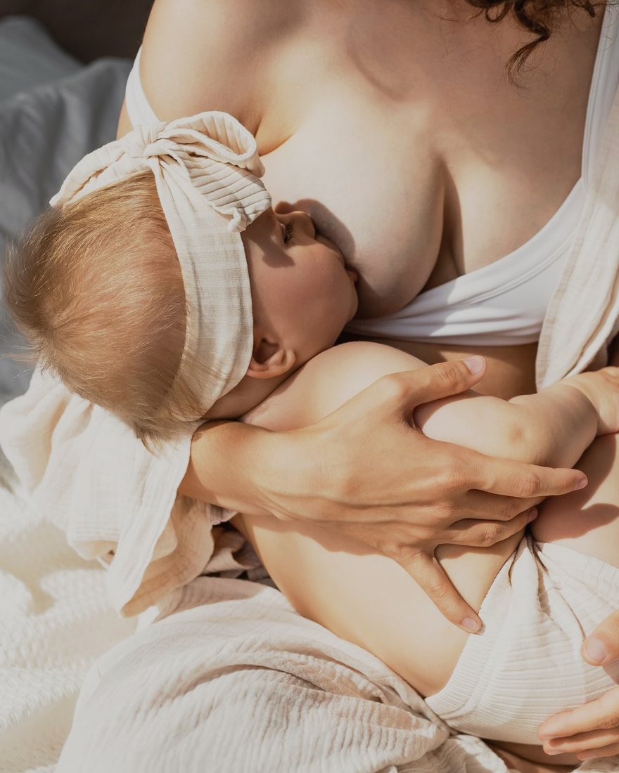 A parent breastfeeding their baby on a bed in warm light, one hand resting on the baby