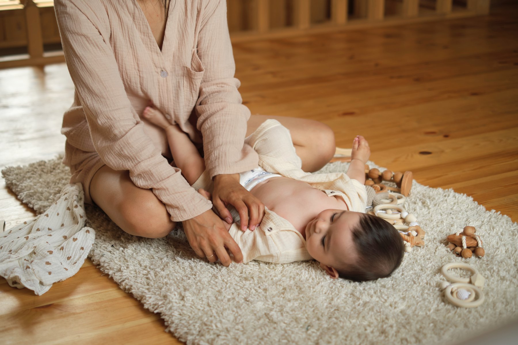 A parent kneeling beside a baby lying on a rug with wooden teething toys nearby