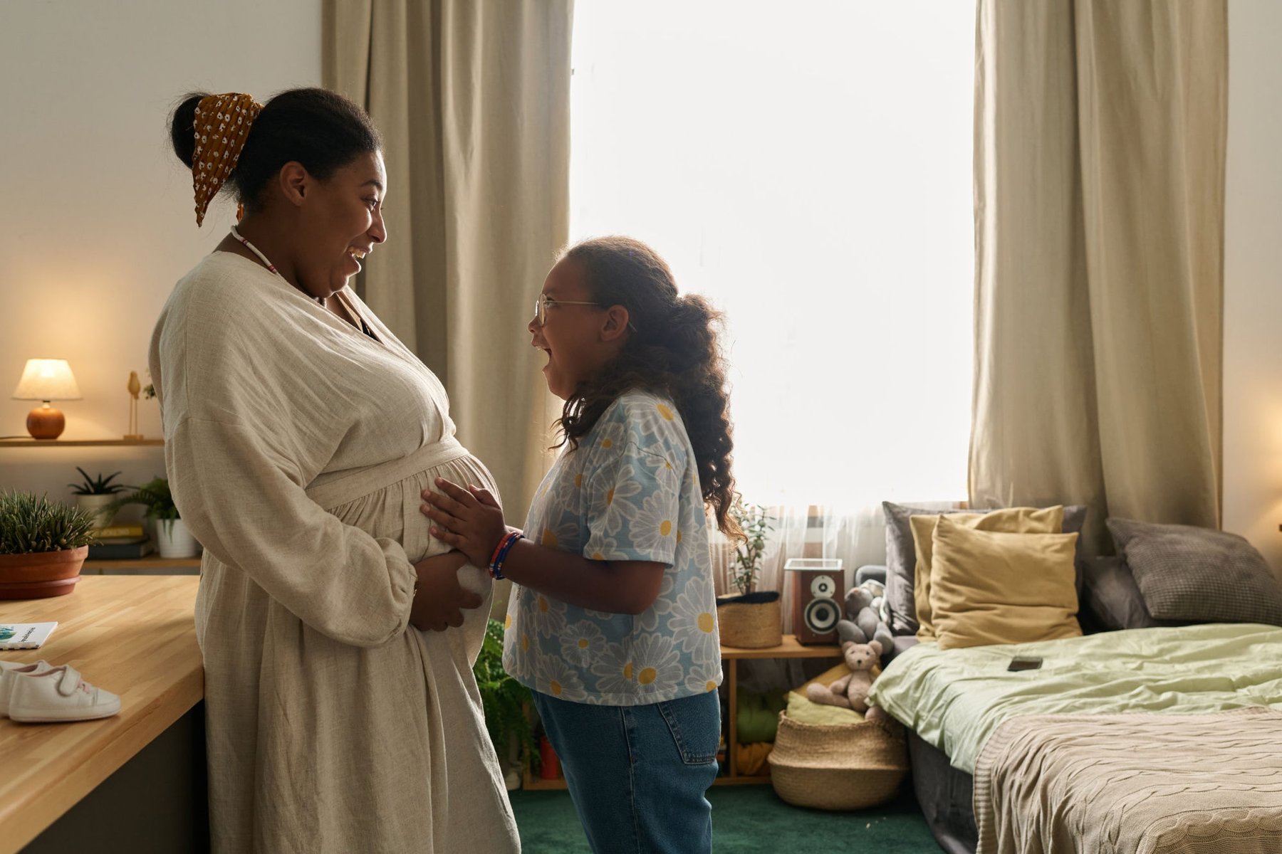 An excited child gently touching a pregnant woman's belly in a sunlit bedroom