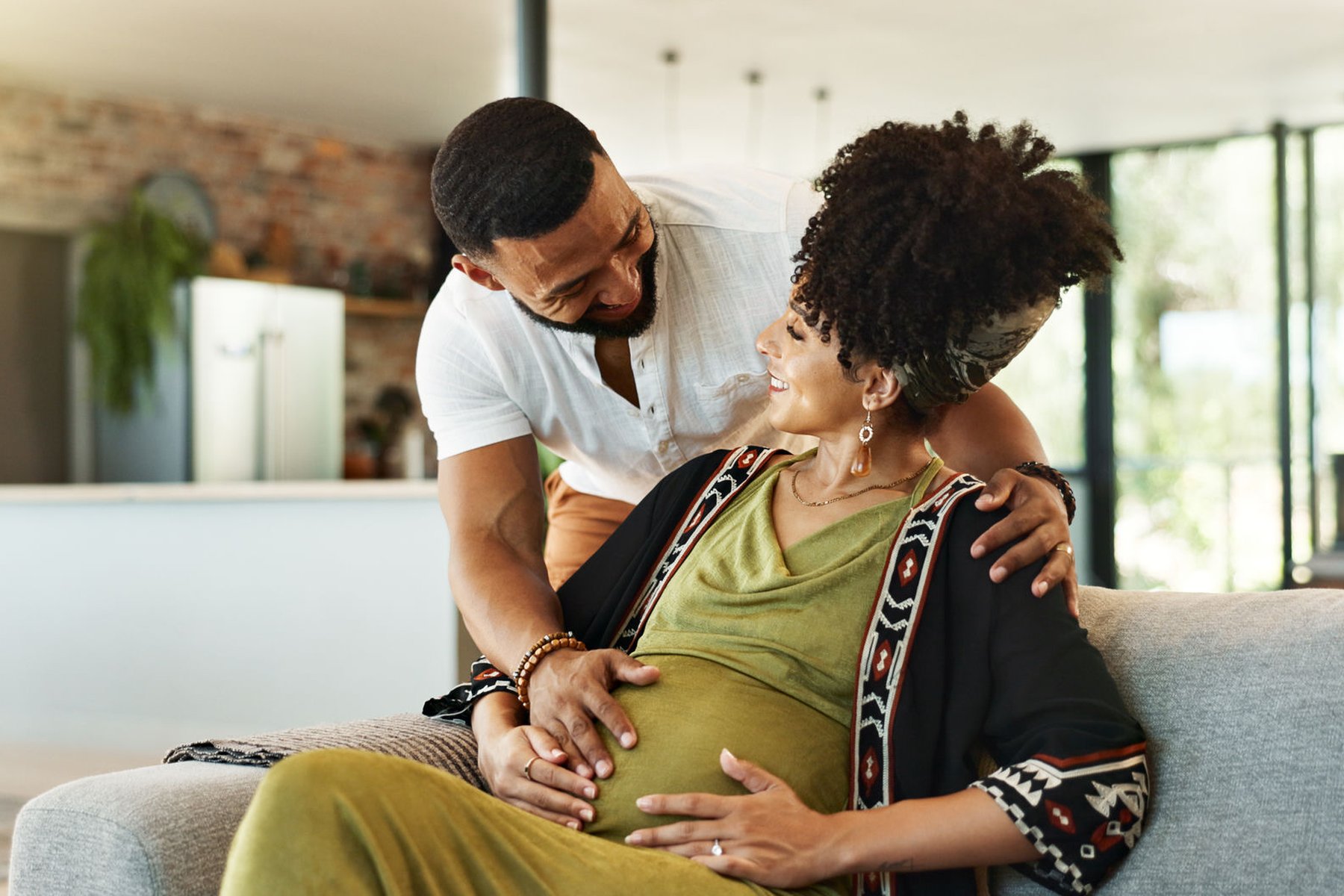 A couple smiling together on a couch, hands resting on the pregnant belly