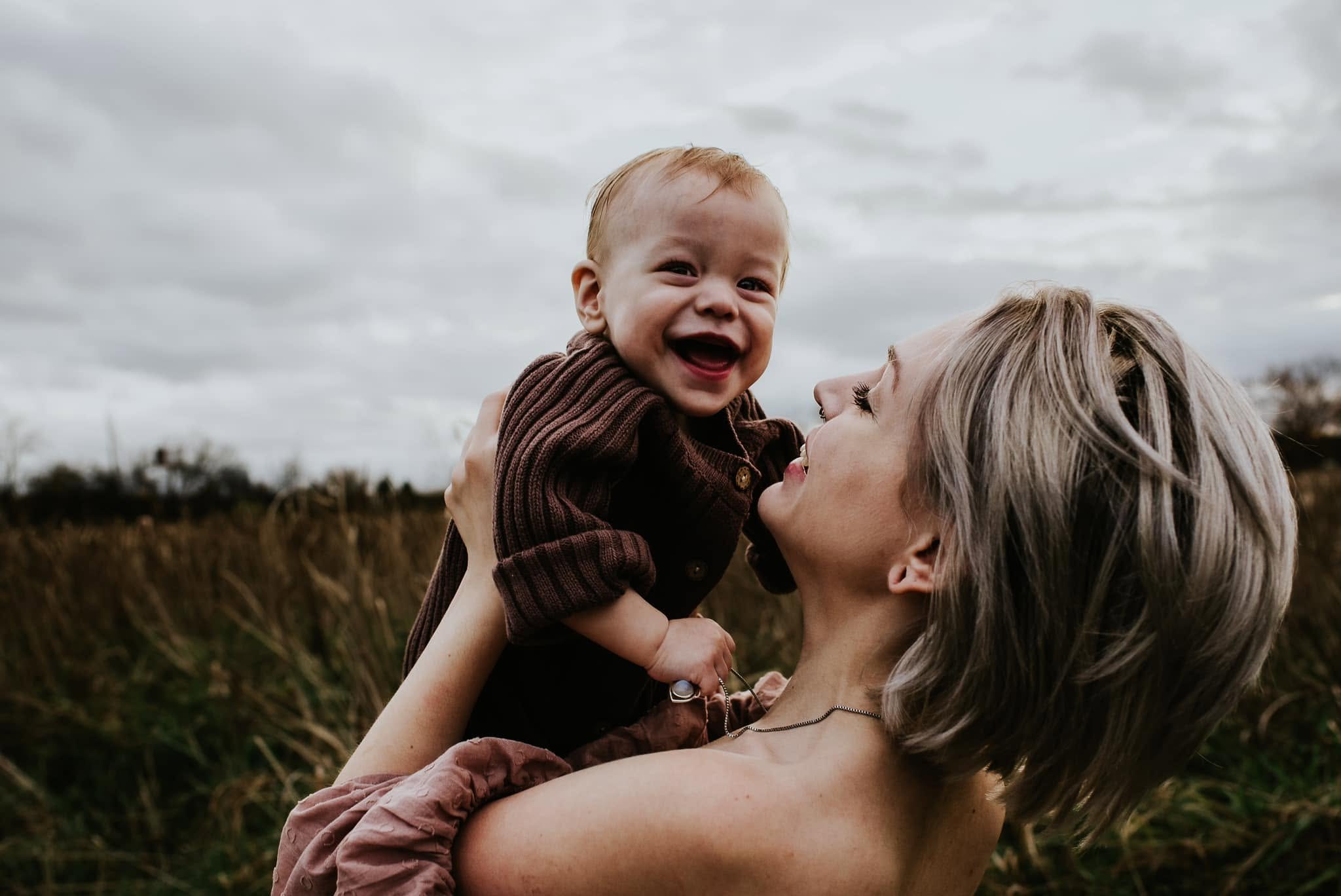 Mady, the doula behind Mycelia Birth Co., lifting her laughing baby son above her in a field under a gray sky