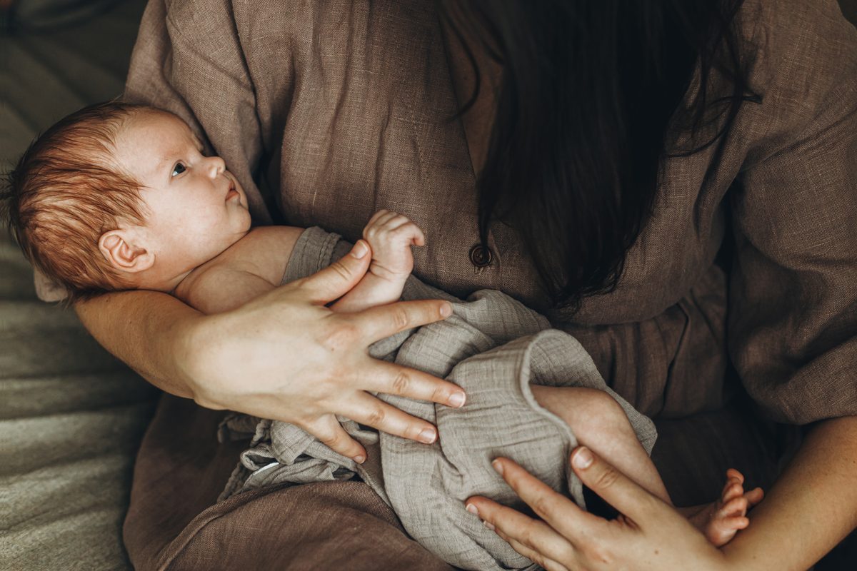 A mother in a linen robe smiling down at her swaddled newborn