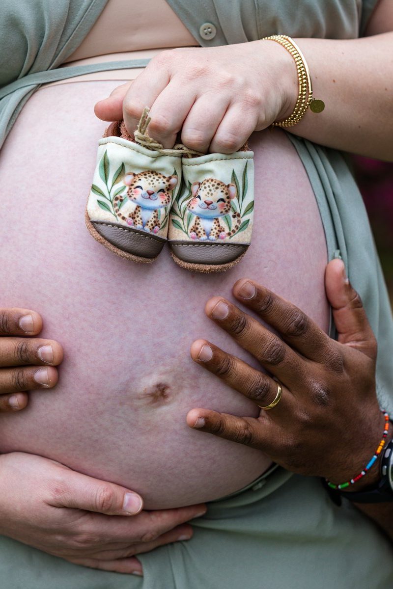 A pregnant belly with hands from both partners, one holding tiny leopard-print baby shoes
