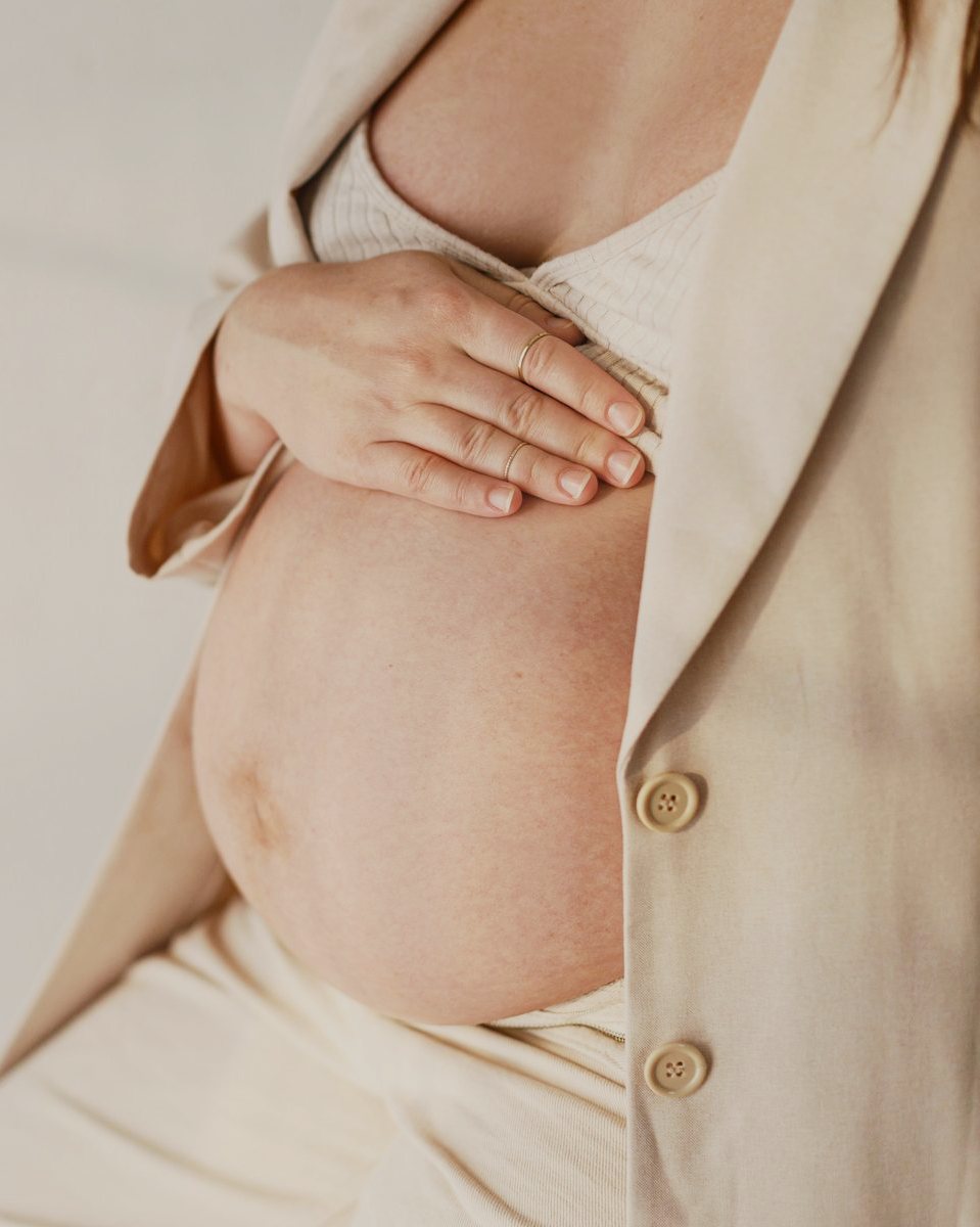 A pregnant person in a cream cardigan resting one hand on their belly in soft window light